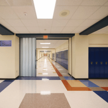 Empty school hallway with blue lockers on both sides, a tile floor with colored patterns, fluorescent ceiling lights, and commercial doors at the entrance—ideal for easy access or automatic door service needs.