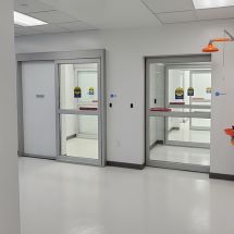 A clean, empty hospital corridor with white walls and floor is lined by commercial doors. Two sleek automatic sliding glass doors are on the left and right, while an orange emergency shower is partially visible on the right.