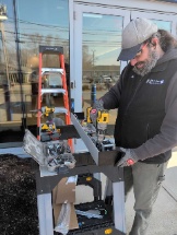 A person wearing a cap and gloves uses a power tool on a metal frame outdoors, possibly performing automatic door repair, with a stepladder and equipment nearby.