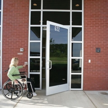 A person in a wheelchair approaches a red brick building, pressing an automatic door service button near the sleek glass entry door.