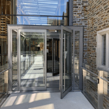 A modern glass door is propped open in a corridor with glass walls and a stone brick exterior visible, highlighting the need for reliable commercial door repair or automatic door service to ensure smooth functionality and security.