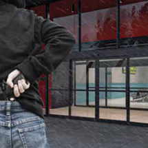 A person in a black hoodie holds a handgun behind their back while standing outside a building with glass commercial doors, possibly awaiting automatic door maintenance or commercial door installation services.