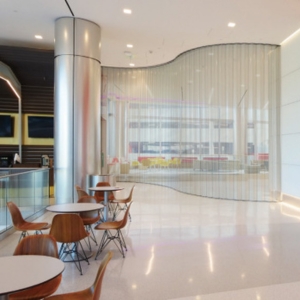 Modern cafe interior with round tables, wooden chairs, and a curved glass partition. The space features sleek automatic door service, seamlessly blending convenience and design.