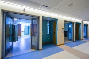 A hospital hallway with open doors leading to individual patient rooms, each partially enclosed by blue privacy curtains. The floors and walls are in neutral tones, complemented by automatic door maintenance ensuring smooth, quiet access for staff and patients alike.