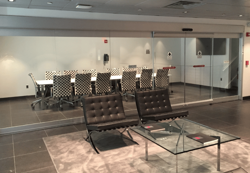 Modern office lounge with two black chairs and a glass coffee table in the foreground, and a glass-walled conference room with checkered chairs and commercial doors in the background.