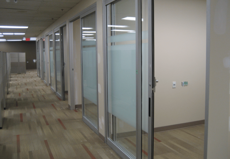 Hallway in an office with glass-walled private offices on the right and cubicles on the left, featuring beige walls and carpeted floors—ideal for businesses needing commercial door repair or automatic door maintenance.